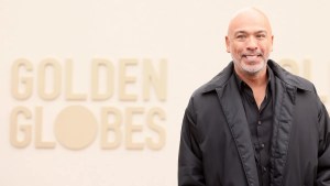 A man wearing a black jacket and shirt with medium-toned skin smiles in front of a beige backdrop with the words "GOLDEN GLOBES" on it.