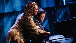 Two Black women at a piano, one leaning over the other's shoulder and helping instruct her as the younger woman looks uncomfortable. They are on stage, the background various shades of dim blue.